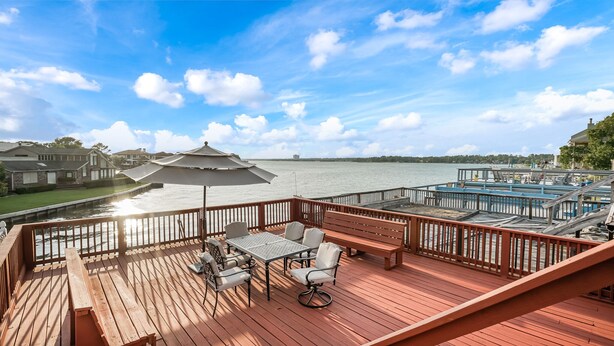 A wooden deck with outdoor furniture, a table, and an umbrella, overlooking a body of water and a dock area.