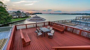 Eine Holzterrasse mit Tisch und Stühlen, mit Blick auf ein Gewässer, im Hintergrund ein Sonnenuntergang.