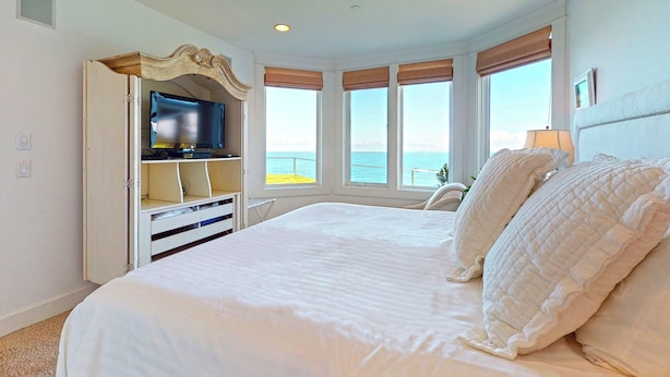 Ein Schlafzimmer mit einem großen Bett, einem Fernseher auf einer Holzkommode und Meerblick durch große Fenster.