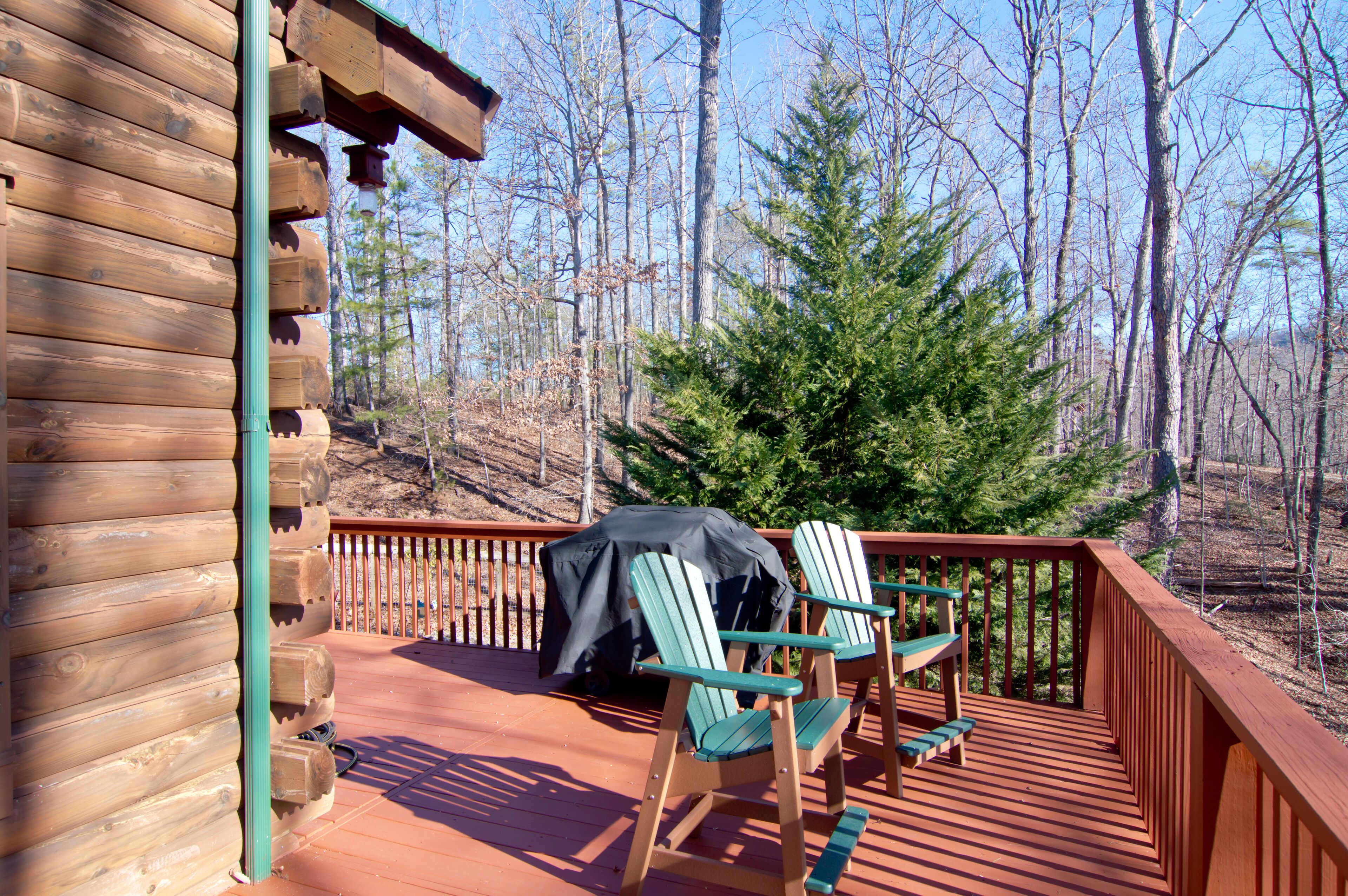 A wooden deck with two Adirondack chairs, a covered grill, and a view of a forest.