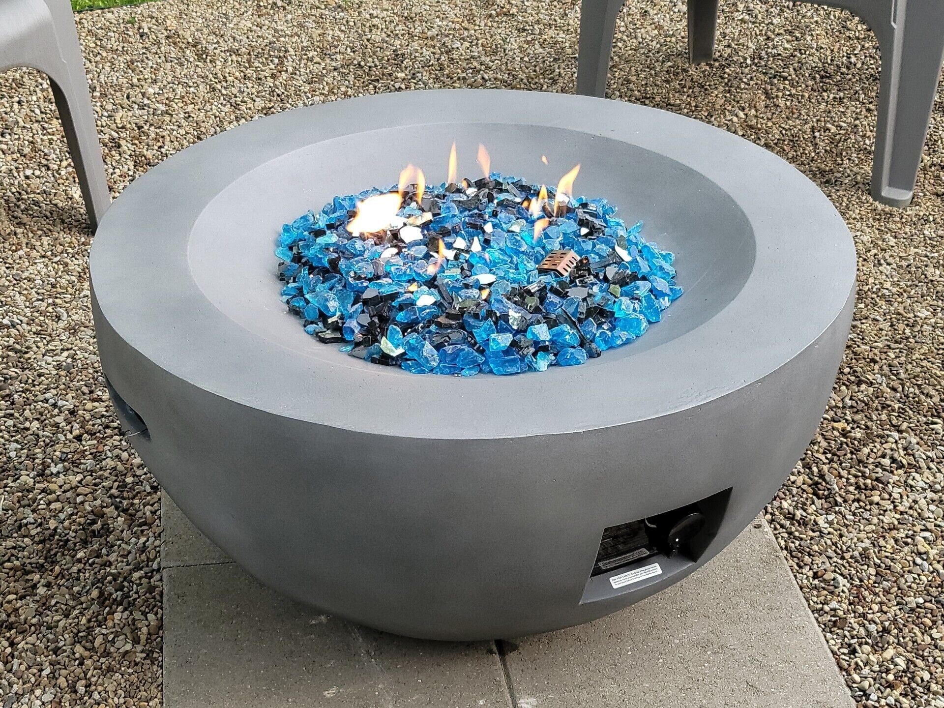 A circular outdoor fire pit with a blue glass top, surrounded by gravel.