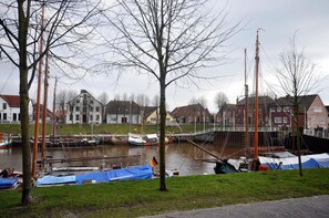 A riverside view with boats docked, a building with a red roof, and a grassy area with trees. - Ferienwohnung Am Hafen West "schoner" (Wittmund)