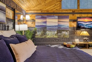 A bedroom with a bed, pillows, a nightstand with a lamp, a TV, and a wall with framed pictures.
