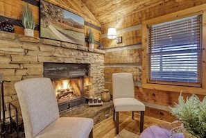 Une pièce chaleureuse avec une cheminée en pierre, des murs en bois et une grande fenêtre équipée de stores.