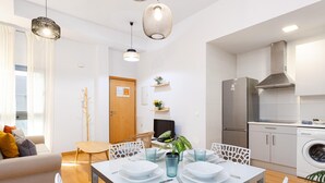A modern kitchen with a stainless steel refrigerator, a built-in oven, and a washing machine. There is a dining area with a white table and chairs, and a sofa in the living room.
