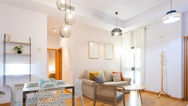 A modern living room with a wooden floor, a sofa, a dining table with chairs, and pendant lights.