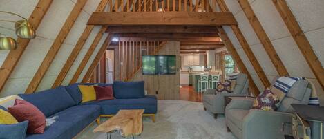 A cozy attic living space with a wooden ceiling, a large sofa, a small table, and a television.
