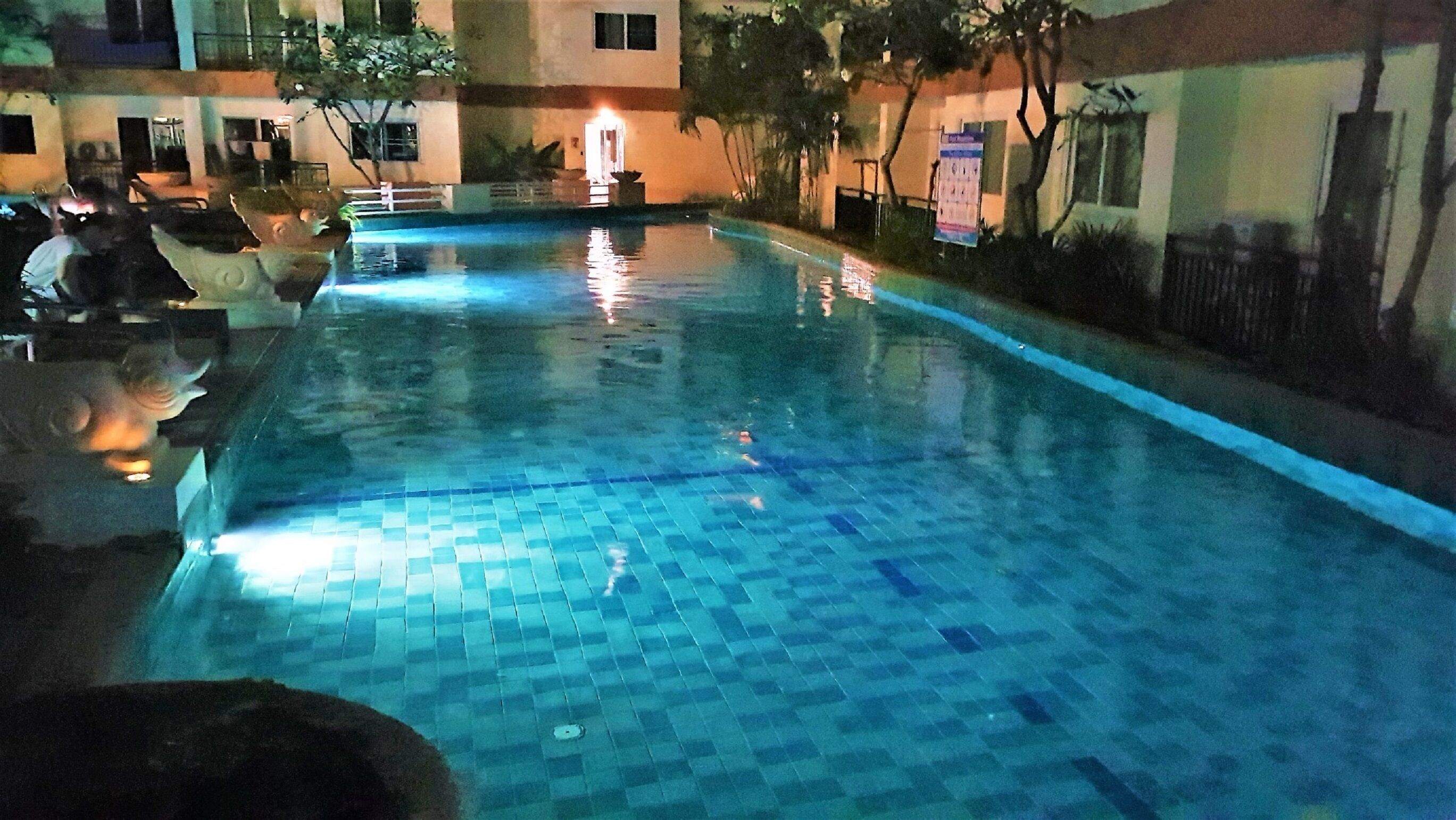 A swimming pool with tiled flooring, surrounded by buildings and trees.