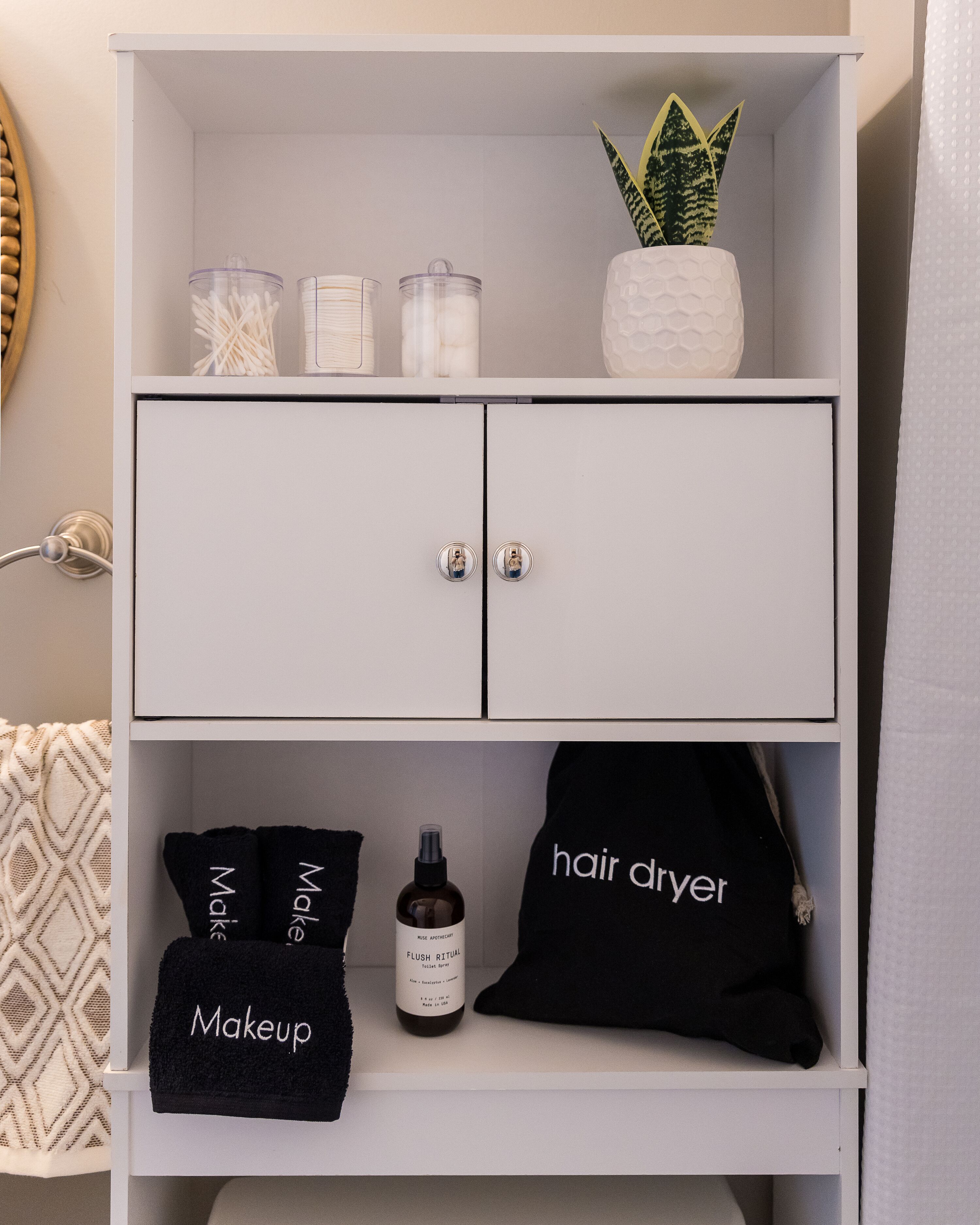 Un meuble blanc à deux portes, contenant un sèche-cheveux, des serviettes pour le maquillage et une bouteille de shampoing.