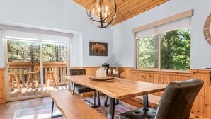 Een houten eettafel met banken, een kroonluchter en een groot raam met uitzicht op een bos.