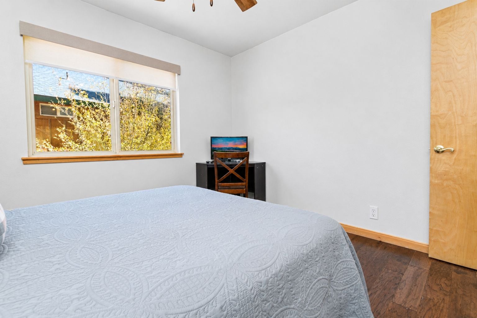 A bedroom with a bed, a TV on a stand, a window with a view of trees, and a wooden door.