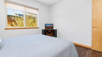 A bedroom with a bed, a TV on a stand, a window with a view of trees, and a wooden door.