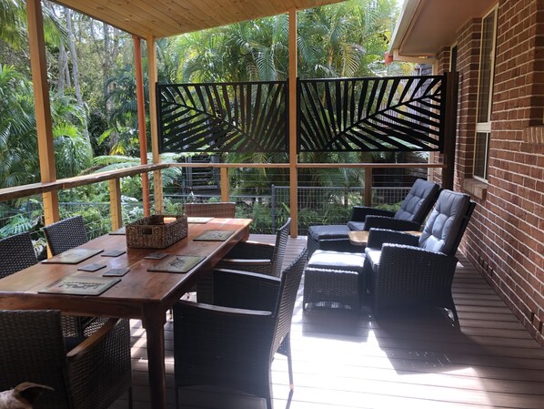 Un espace extérieur aménagé, abrité et doté de mobilier en osier et d’une table en bois, le tout entouré d’une végétation luxuriante.