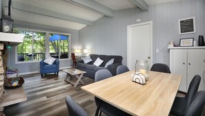 A modern living room with a wooden dining table, a blue armchair, and a grey sofa. There is a large window with a view of greenery outside. - Beautiful 2-bedroom Cottage (Waupaca)