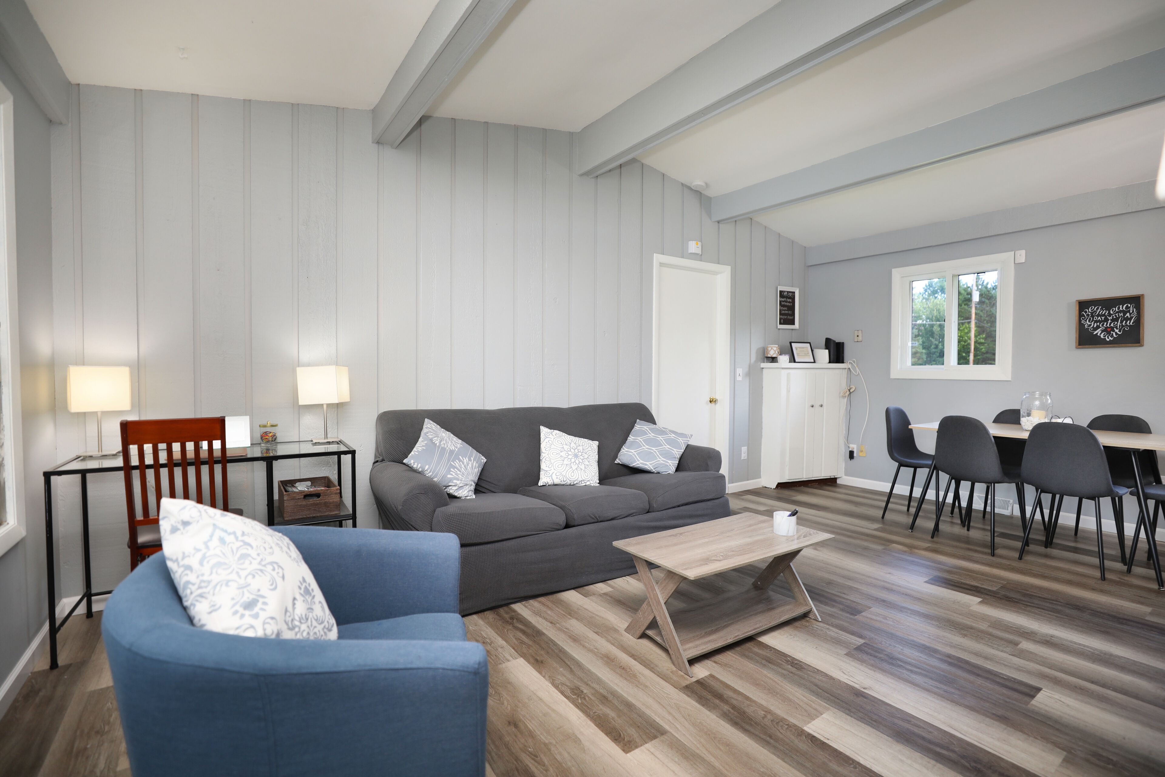 A living room with a grey sofa, blue armchair, wooden coffee table, and a dining area with a table and chairs.
