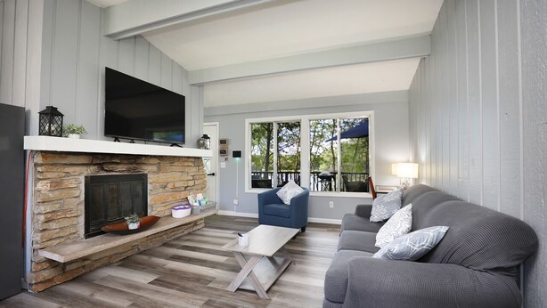 A living room with a stone fireplace, a flat-screen TV, a grey sofa, and a dining area with a table and chairs. - Beautiful 2-bedroom Cottage (Waupaca)