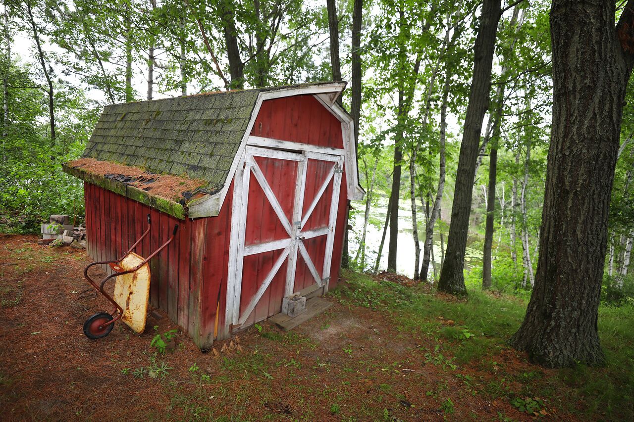 A small, rustic wooden shed with a sloped roof, situated in a forested area near a body of water.