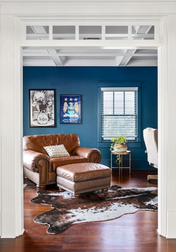 A room with a brown leather sofa, a small round table with a plant, a blue wall, and a window with blinds. - Luxury 6-Bedroom Home in Prime Decatur Neighborhood, Near DT ATL (Decatur)