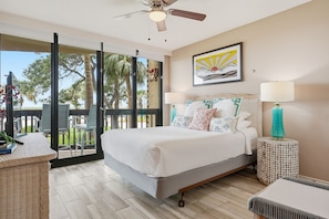 A bedroom with a bed, two bedside lamps, a ceiling fan, a window with a view of trees, and a framed picture on the wall.