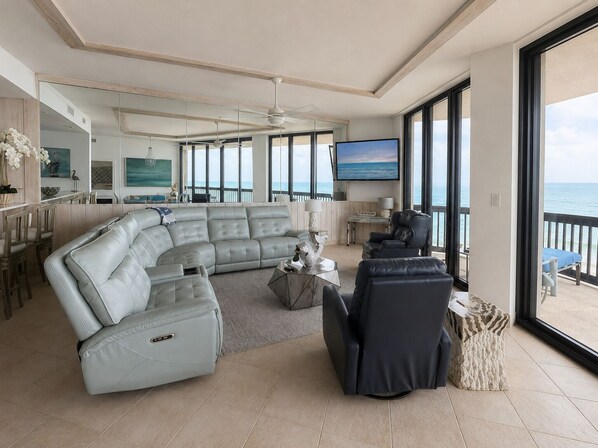 A spacious living area with a large sectional sofa, a black armchair, and a coffee table. There is a television mounted on the wall, and the room has large windows offering a view of the ocean. - Sandpiper Condominiums 1206, Panoramic Gulf Views w/ direct access to Mustang Island/Port A beaches. (Port Aransas)