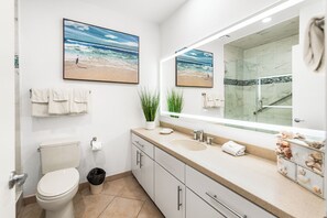 Un baño con inodoro, lavamanos y ducha. Hay dos cuadros con imágenes de una playa en la pared.