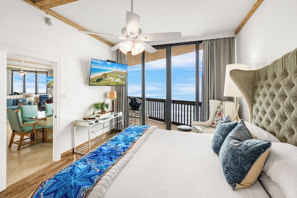 A bedroom with a large bed, a ceiling fan, a TV, and a balcony with a view.