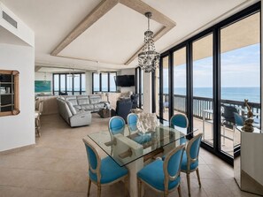 A spacious living area with a glass dining table, blue chairs, a chandelier, and a view of the ocean. - Sandpiper Condominiums 1206, Panoramic Gulf Views w/ direct access to Mustang Island/Port A beaches. (Port Aransas)