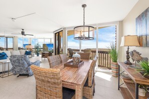 A spacious living area with a large wooden dining table, wicker furniture, and a view of the ocean.