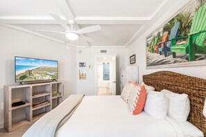Quarto com uma cama grande, um ventilador de teto, uma TV em uma prateleira de madeira e uma pintura com tema praiano na parede.