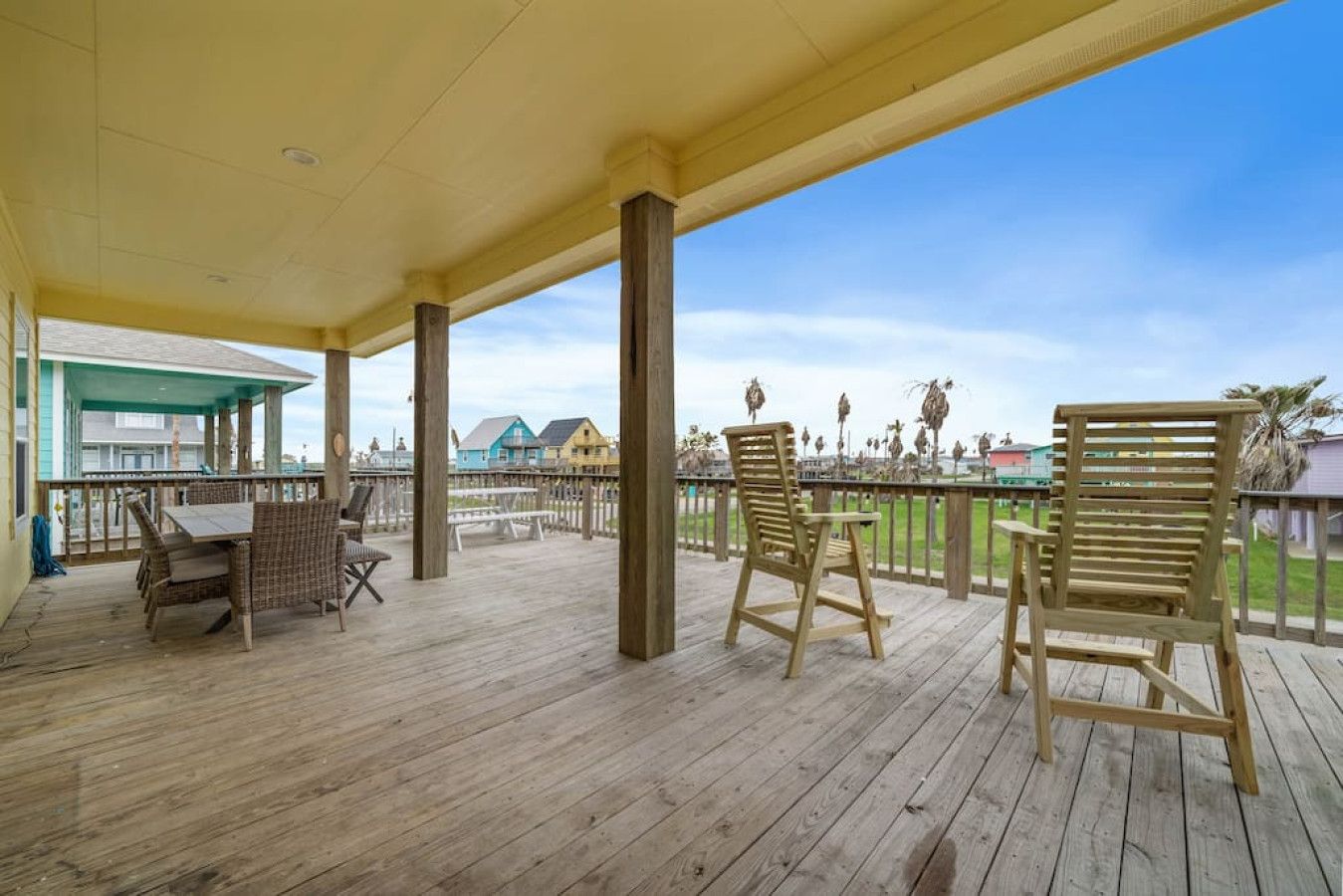 A wooden deck with two wicker chairs and a table, overlooking a beach with palm trees and houses.