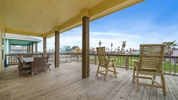 A wooden deck with two wicker chairs and a table, overlooking a beach with palm trees and houses.