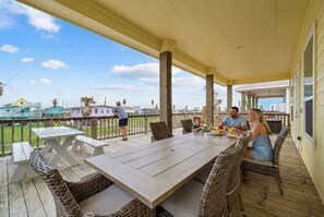 Un patio techado al aire libre con una mesa grande y sillas de mimbre, con vista a una zona residencial con casas y cielo despejado.