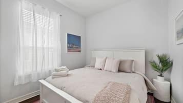 Een slaapkamer met een bed, een raam met dunne gordijnen, een lijst met een afbeelding aan de muur, en een kleine plant op een nachtkastje.