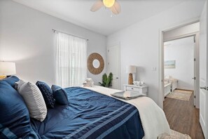 Un dormitorio con una cama grande, una mesita de noche con lámpara, un espejo redondo en la pared y una puerta que da a otra habitación.