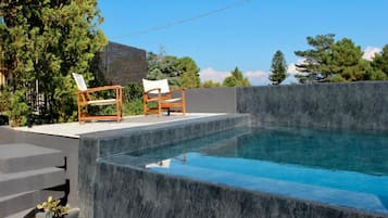 A modern outdoor swimming pool with a stone wall, two wooden chairs, and a wooden deck.