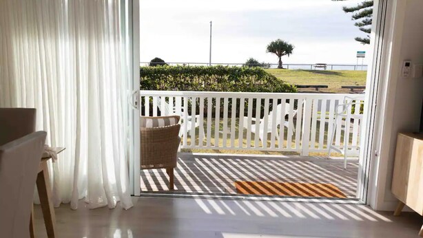 Una habitación con puerta de vidrio corredera que da a un balcón al aire libre con valla blanca, una silla de mimbre y una alfombra pequeña.