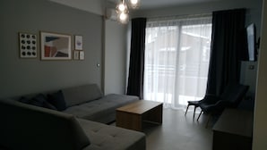 A modern living room with a grey sofa, a wooden coffee table, and a black armchair. There are framed artworks on the wall and a television on the right.