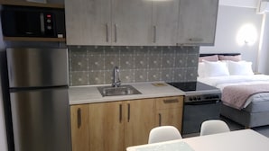 A compact kitchen with a stainless steel refrigerator, a microwave, and a sink. There is a bed with a white and pink pillow arrangement.