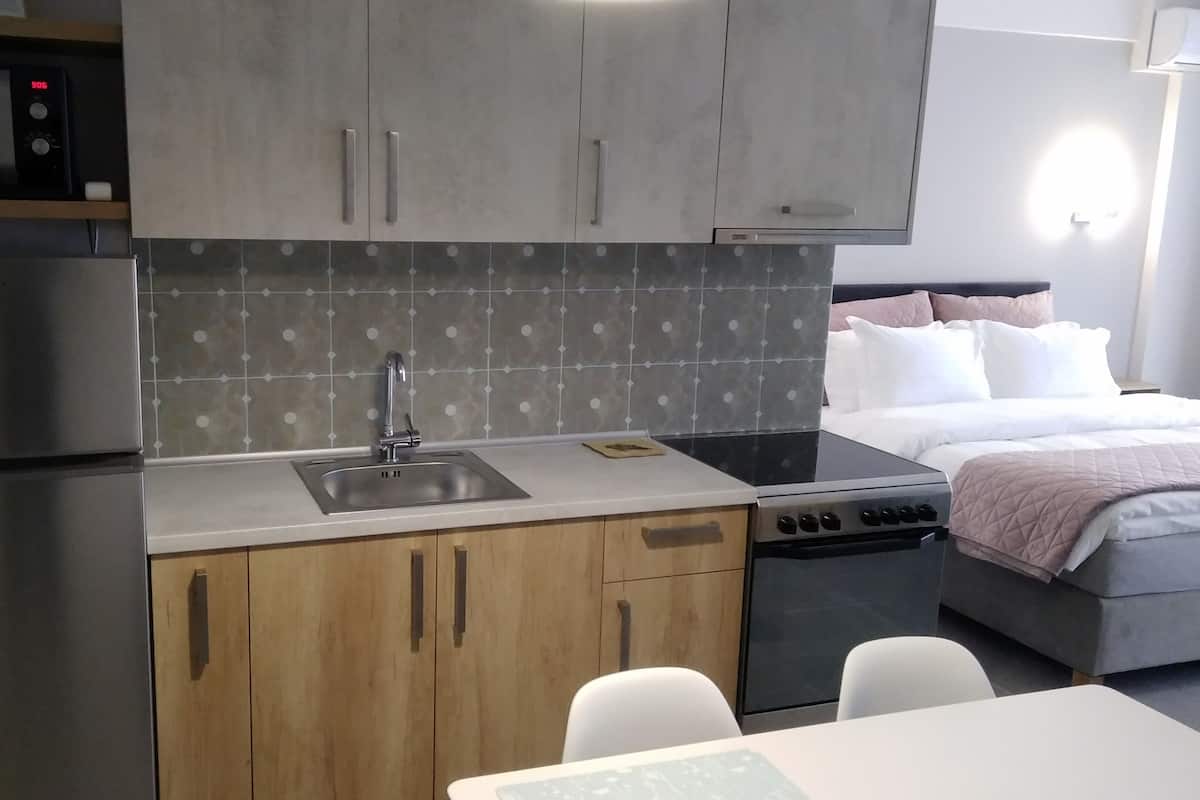 Cocina compacta con refrigerador de acero inoxidable, microondas y pileta. Se puede ver una cama con almohadones blancos y rosados.