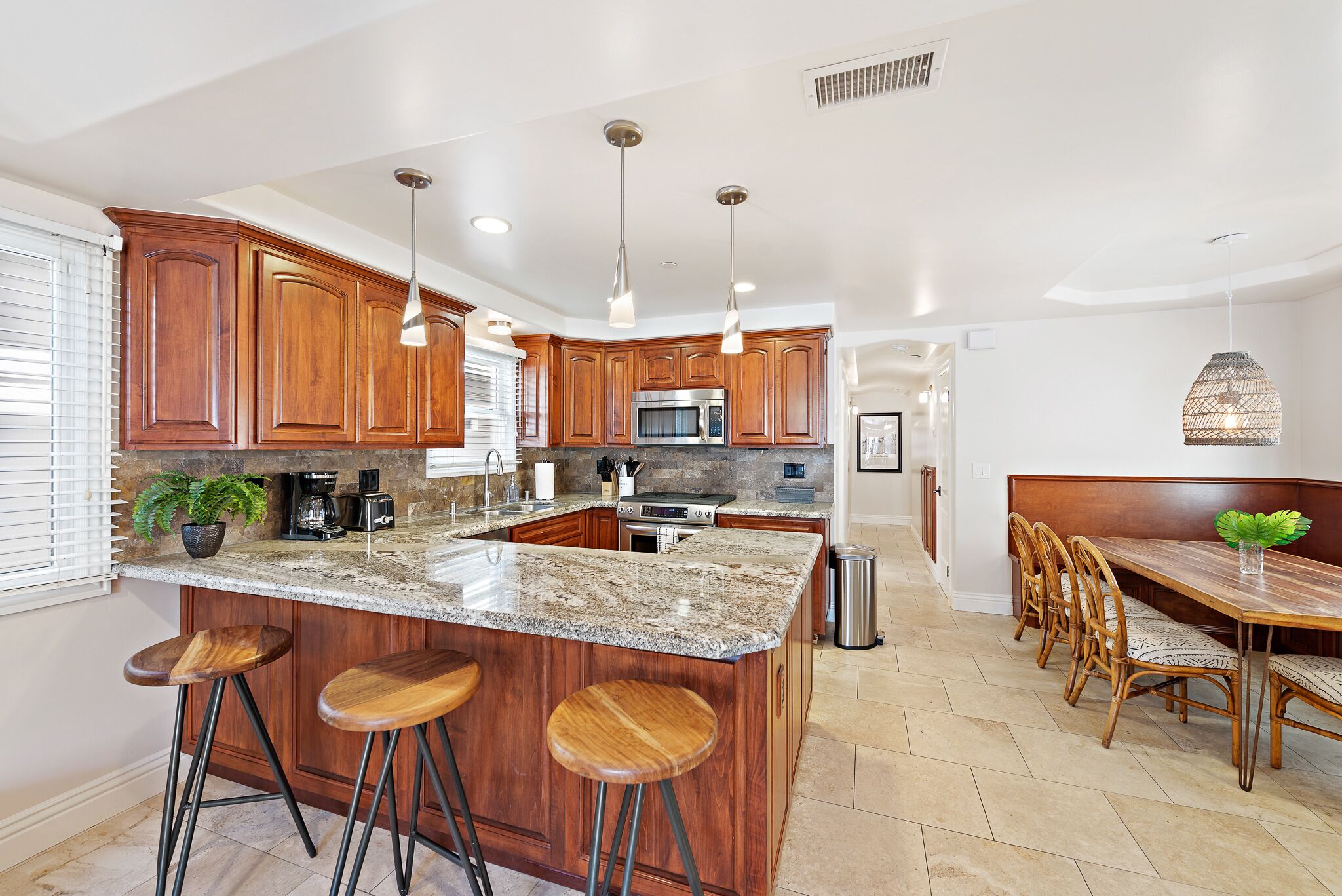 A kitchen with wooden cabinets, a granite countertop, a central island with stools, a dining area with a bench, and pendant lighting.
