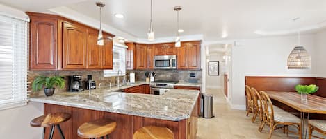 A kitchen with wooden cabinets, a granite countertop, a central island with stools, a dining area with a bench, and pendant lighting.