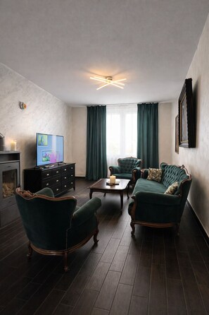 Una sala de estar con piso de madera oscura, una chimenea, un televisor, una mesa de centro y dos sofás tapizados en color verde.