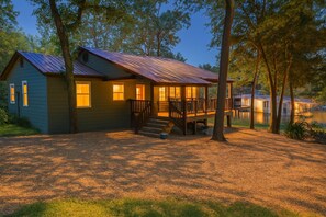 Une cabane dotée d’un porche et d’une terrasse, entourée d’arbres et bordée par un plan d’eau.