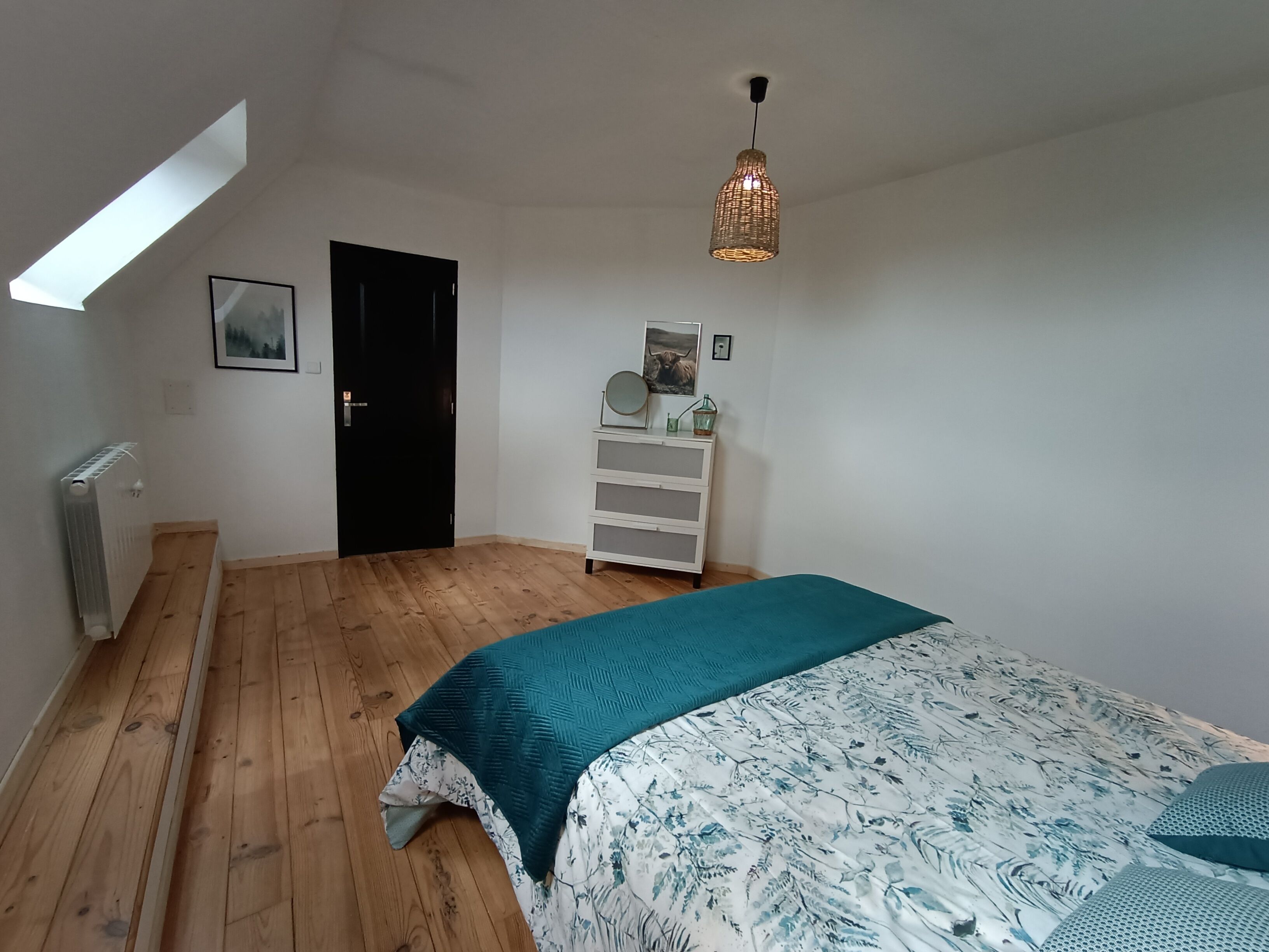 Ein Schlafzimmer mit Bett, Kommode, Heizkörper, Tür und Dachfenster.