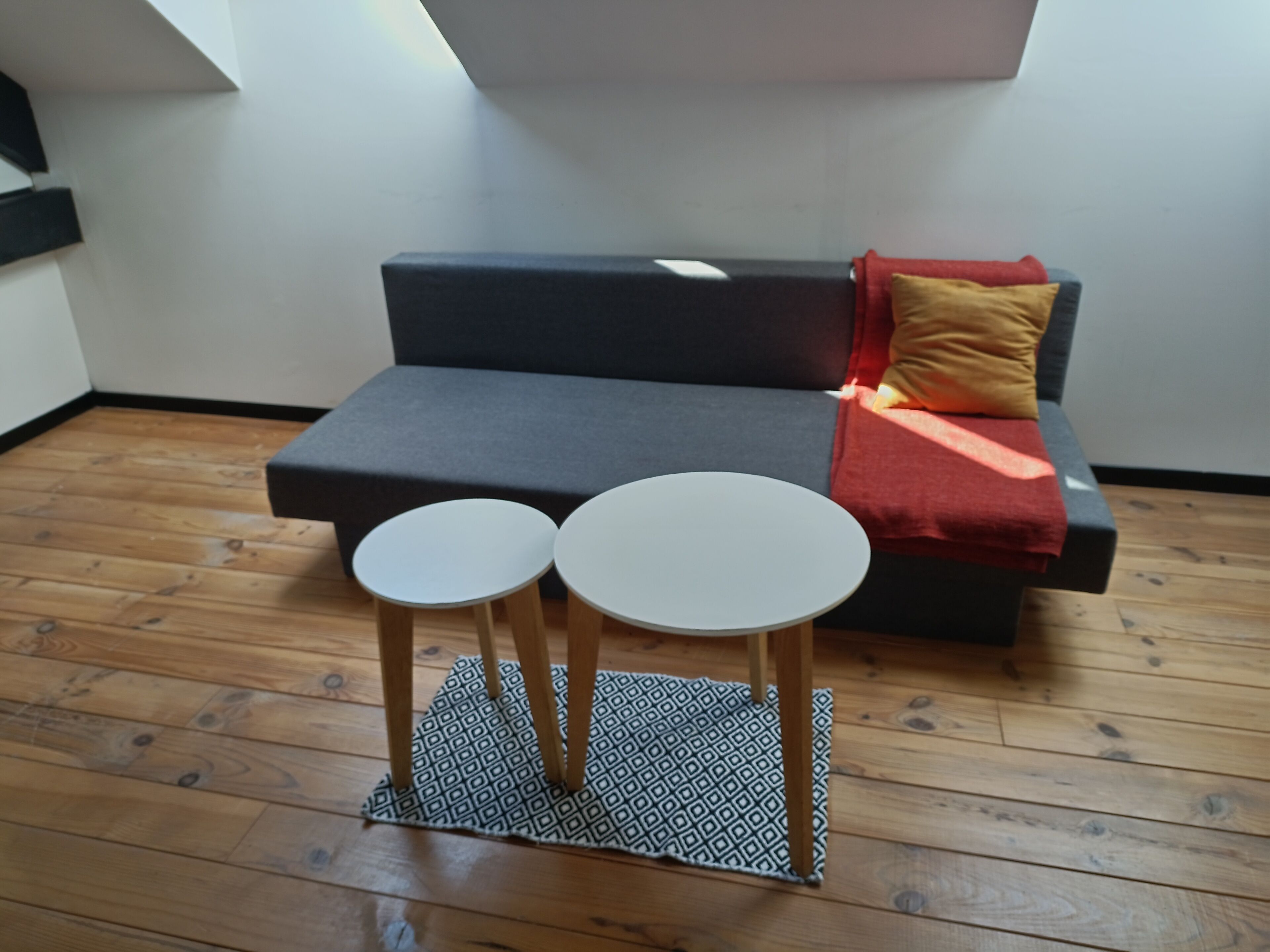 Ein modernes Wohnzimmer mit einem grauen Sofa, zwei runden Tischen und einem gemusterten Teppich auf Holzdielen.