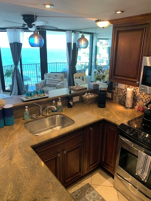 A kitchen with a large window, a sink, a stove, and a dining area with a view of the sea.