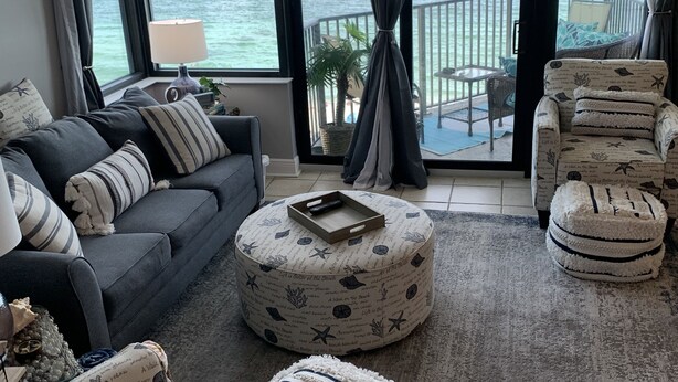 Una sala de estar con sofá, reposapiés y sillón, una mesita y vista al océano a través de amplias ventanas.