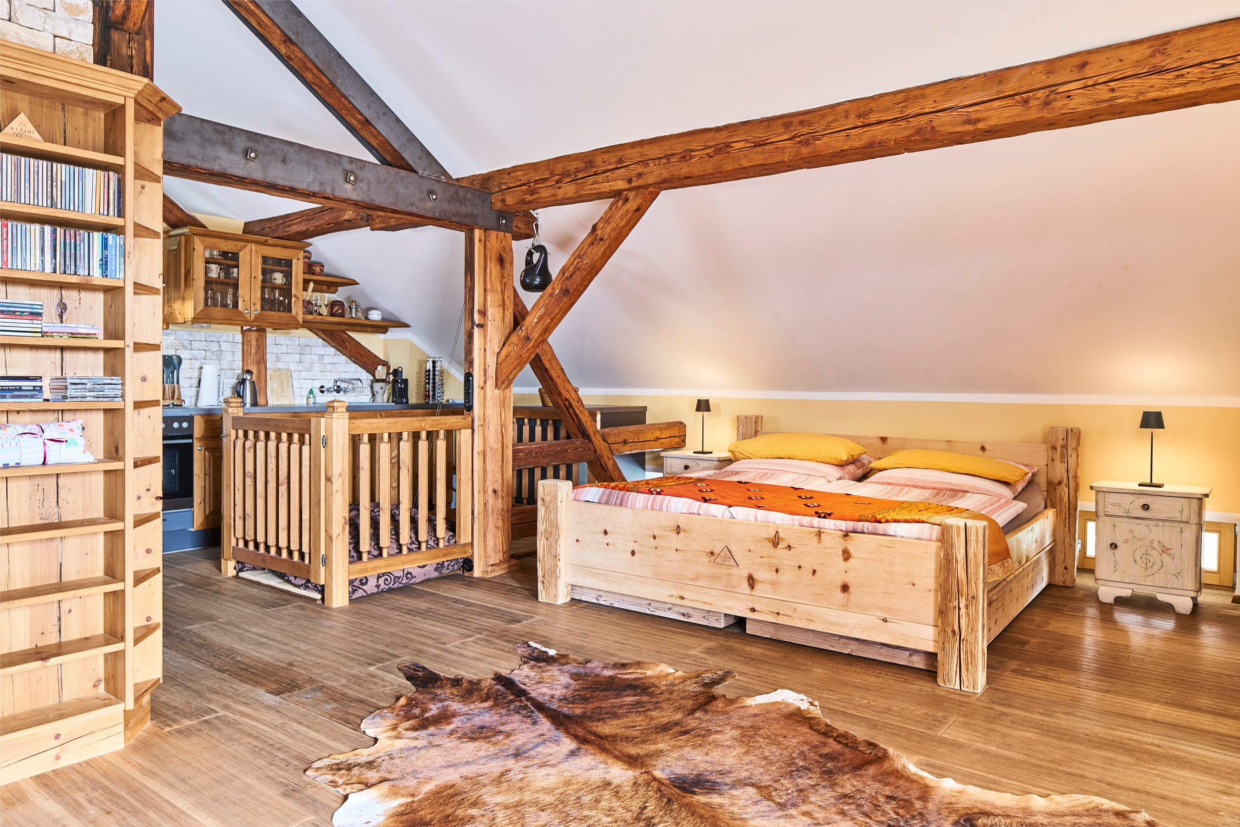 Une chambre à coucher rustique avec un plafond en pente, des poutres en bois, un lit recouvert d’une couvre-lit à motifs, une bibliothèque et un tapis en cuir.