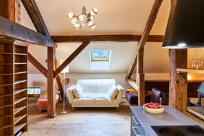 An attic room with exposed wooden beams, a white sofa, a dining table with a bowl of apples, and a modern chandelier. - Ferienwohnung mit Balkon und Bergblick (Bad Feilnbach)