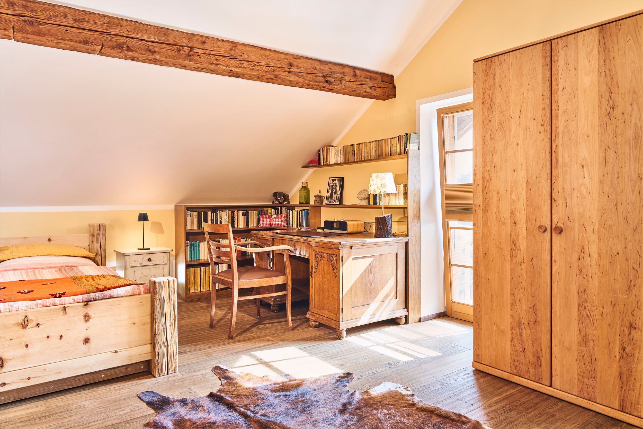 Ein gemütliches Zimmer mit schrägen Wänden, Holzmöbeln, einer Bücherwand und einem großen Kleiderschrank.